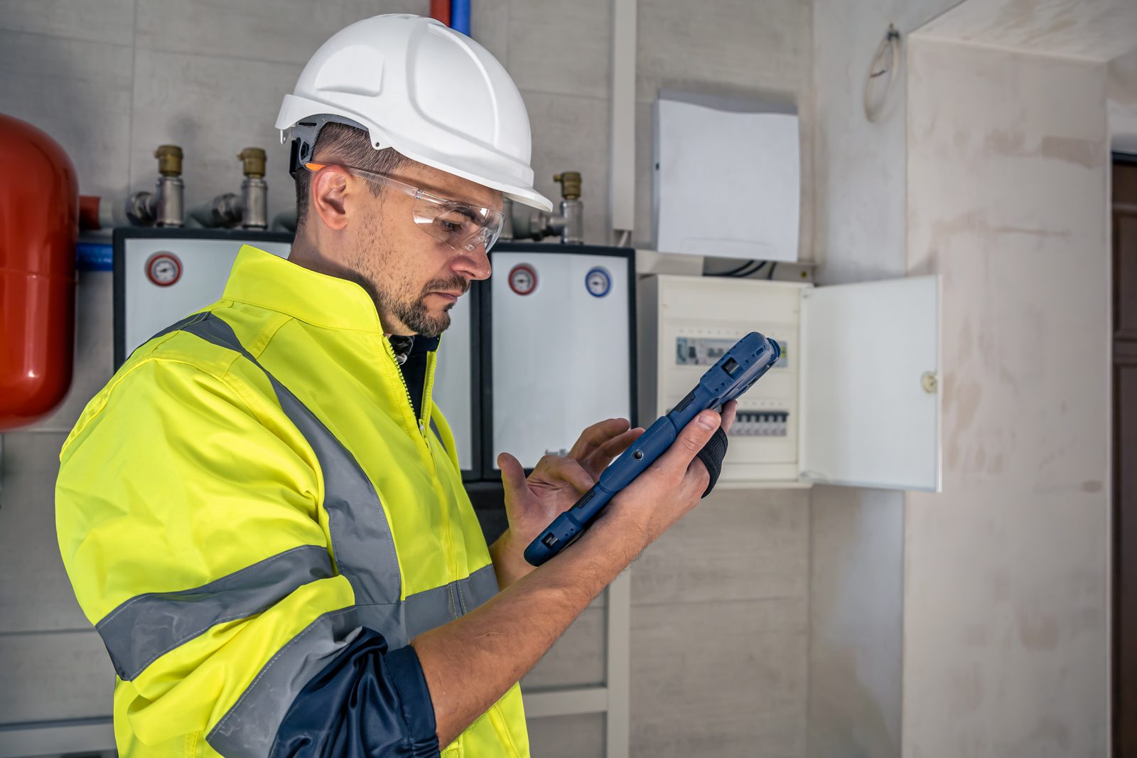 An engineer in high visibility clothing inspecting an electrical panel with a tablet